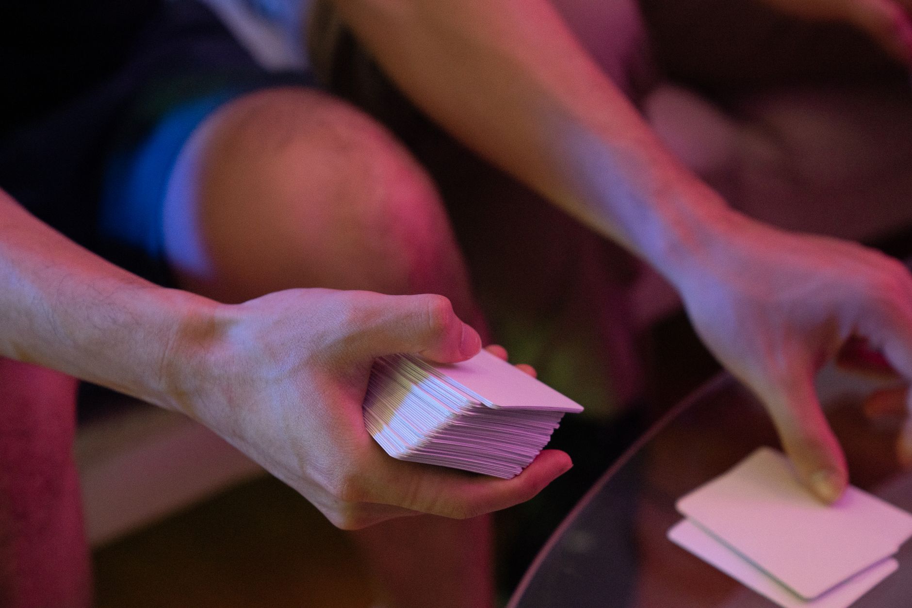 Close-up of hands shuffling a deck of cards on a glass table with pink lighting.